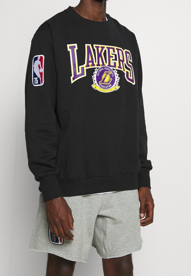 Sudadera negra con "LAKERS" en púrpura y amarillo, logo de la NBA en la manga, cuello redondo y puños y dobladillo acanalados.