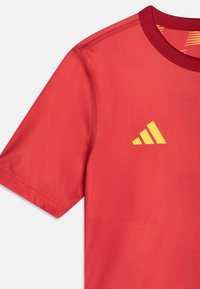 Maglia sportiva rossa con colletto rotondo e maniche corte. Presenta il logo Adidas giallo, una texture liscia e dettagli di cuciture rinforzate.