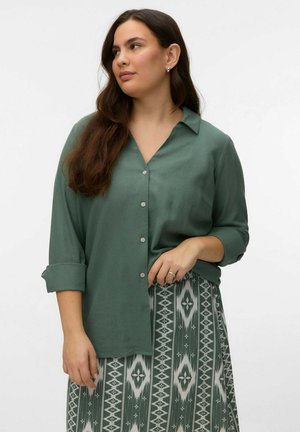 Femme aux longs cheveux bruns portant une chemise verte à boutons et une jupe verte et blanche à motifs, debout devant un fond uni.