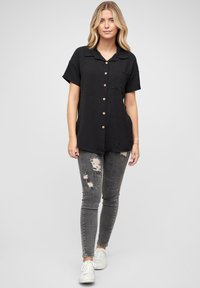 Camicia nera a maniche corte con bottoni e colletto, dotata di una tasca sul petto sinistro e bottoni in legno. Abbinata a jeans skinny grigi strappati e sneakers bianche.