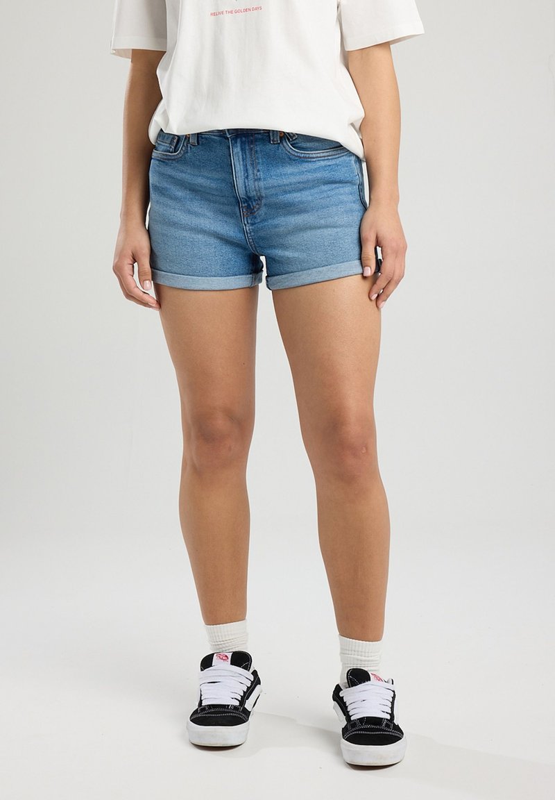 Shorts di denim in azzurro chiaro, orli arrotolati, design a cinque tasche. Abbinati a sneaker nere e calzini bianchi alla caviglia.