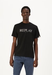 Camiseta de algodón negra con un gran gráfico gris "REPLAY" en la parte frontal. Diseño de cuello redondo con mangas cortas. Combinada con jeans de color azul claro.