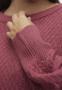 Pull en tricot rose poudré, présentant un motif texturé, des poignets côtelés et un col rond avec une finition douce et confortable.