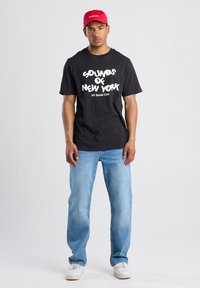 Black cotton t-shirt featuring white text "SOUNDS OF NEW YORK" and "NY Social Club." Worn with light blue jeans and a red cap.