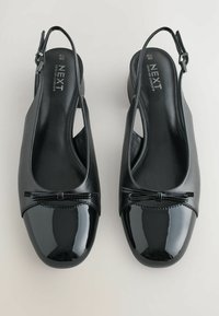 Juodos slingback bateliai su blizgiu lakuotu kojų pirštu ir kaspino detale, pagaminti iš lygaus odos. Jie turi atvirą kulną ir žemo profilio dizainą.