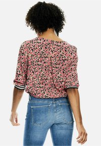 Kobieta o kręconych włosach, ubrana w różową, kwiatową bluzkę z czarno-białymi mankietami i niebieskie dżinsy, stoi tyłem na białym tle.