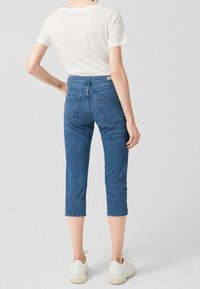 Pantalones capri de mezclilla azul con una cintura ajustada, dos bolsillos traseros, longitud recortada y una textura suave, combinados con una camiseta blanca y zapatillas deportivas.