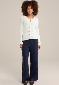 Bela striped cardigan s sprednjim gumbom, dvema žepoma in rebrastimi manšetami, v kombinaciji z mornarsko širokimi hlačami in rjavimi pikastimi čevlji.