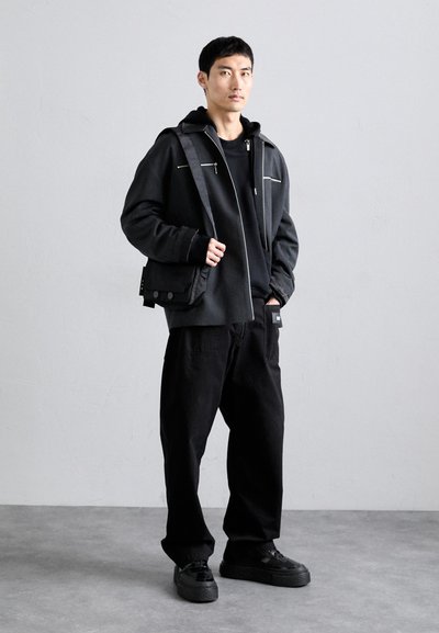 Jaqueta preta com piping branco, moletom preto por baixo, combinada com calças largas pretas e ténis pretos texturizados. Design simples e moderno.