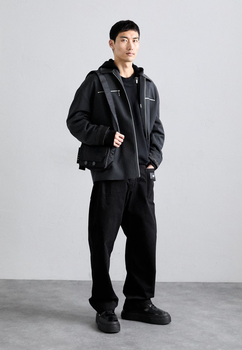 Veste noire avec des passepoils blancs, un sweat à capuche noir en dessous, associée à un pantalon noir ample et des baskets noires texturées. Design simple et moderne.