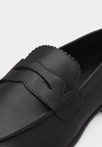 Mocassin noir en cuir avec surpiqûres apparentes sur le dessus, bride en selle avec fente et détail festonné sur le dessus du pied.