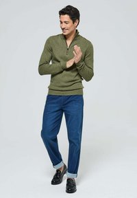 Pull en maille vert olive avec un col zippé, associé à un pantalon bleu foncé à manches retroussées et des mocassins noirs, offrant une coupe décontractée.