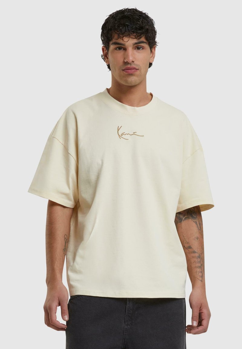 T-shirt surdimensionné crème en coton avec un col rond et des manches courtes. Présente une signature brodée en or sur la poitrine.