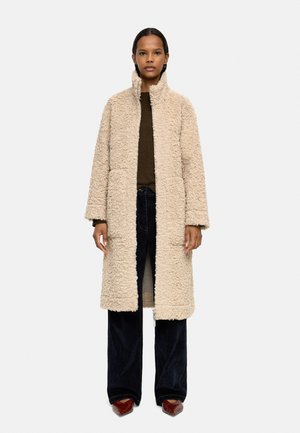 Beżowy płaszcz futrzany z wysokim kołnierzem, strukturalna powierzchnia, dwie przednie kieszenie, noszony na ciemnej bluzce i szerokich dżinsach z czerwono-brązowymi butami.