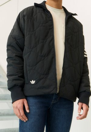 Mężczyzna ubrany w czarną pikowaną kurtkę na zamek marki Adidas z logo i paskami na rękawie, na białą koszulę i niebieskie dżinsy.