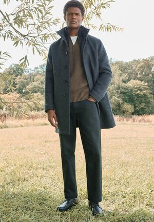 Abrigo de lana gris con cuello alto, jersey de punto marrón debajo, pantalones oscuros y zapatos negros, de pie en un entorno exterior con hierba.