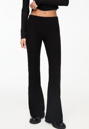 Vrouw draagt hoog getailleerde zwarte gebreide flarebroek en zwarte chunky laarzen, met een zwarte cropped longsleeve top, staand tegen een witte achtergrond.