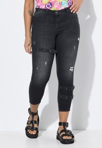 Pantalons capri en denim noir avec des détails déchirés, ornés d'accents en sangle et de quincaillerie en métal. Portés avec des sandales noires à lanières.