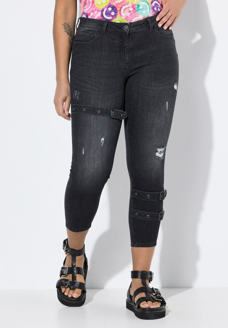 Pantalons capri en denim noir avec des détails déchirés, ornés d'accents en sangle et de quincaillerie en métal. Portés avec des sandales noires à lanières.