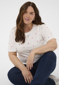 Weißes T-Shirt mit kurzen Ärmeln und verstreuten kleinen roten Herzmustern. Das Modell trägt dunkle Jeans und sitzt entspannt mit einem gelassenen Ausdruck.