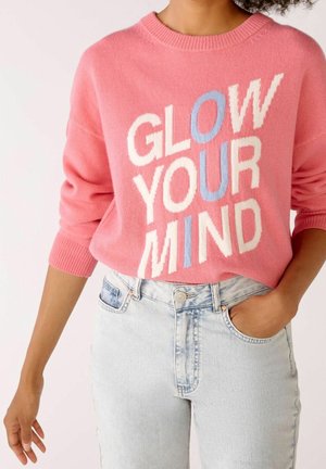 Person iført lyserød sweater med teksten "GLOW YOUR MIND" i hvid og blå, kombineret med lyseblå højttaljede jeans, den ene hånd i lommen.