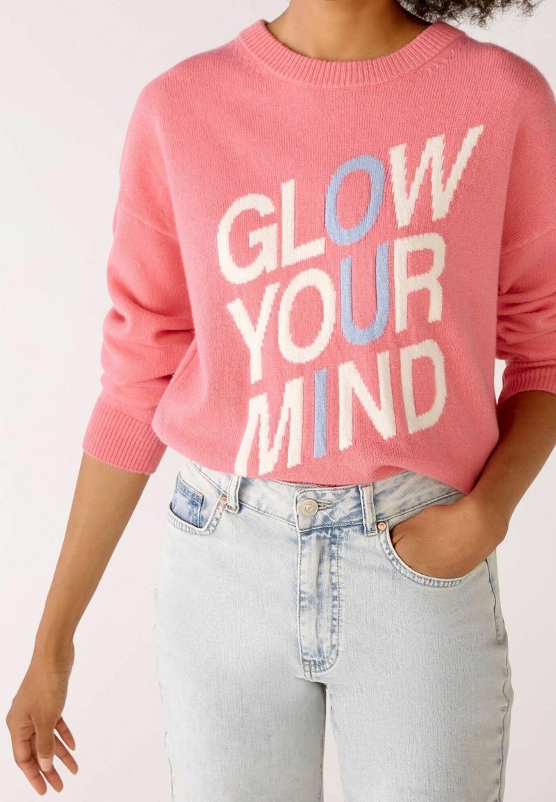 Personne portant un pull rose avec le texte « GLOW YOUR MIND » en blanc et bleu, associé à un jean taille haute bleu clair, une main dans la poche.