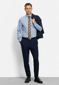 Hellblaues Hemd, braun-weiß gestreifte Krawatte, marineblaue Hose, schwarze Schuhe und dunkelblauer Blazer über der Schulter getragen. Maßgeschneiderte Passform.