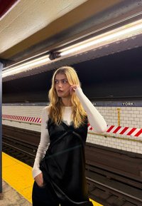 Fekete selymes ruha csipke díszítéssel, fehér hosszú ujjú felső fölött viselve, egy metróállomáson, sárga peronnal és csempézett fallal.