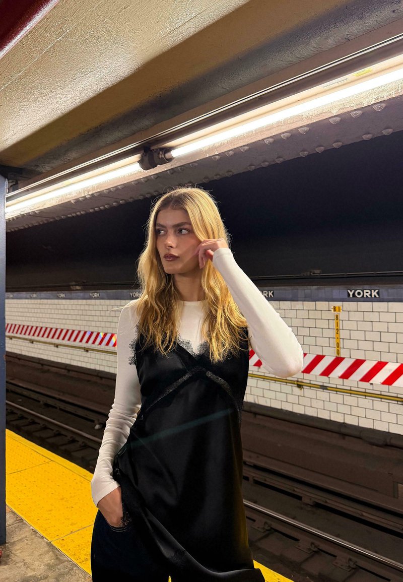 Fekete selymes ruha csipke díszítéssel, fehér hosszú ujjú felső fölött viselve, egy metróállomáson, sárga peronnal és csempézett fallal.