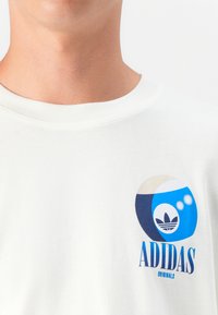 Hvid bomulds t-shirt med et blåt og cremefarvet grafisk design. "ADIDAS" og "ORIGINALS" trykt under et stiliseret logo.