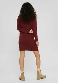 Burgundy pitkähihainen mekko, polvipituinen, sileä kangas. Yhdistetty beigeihin nilkkabootseihin ja kermanvärisiin saapasaitoihin, esitetty takaa.