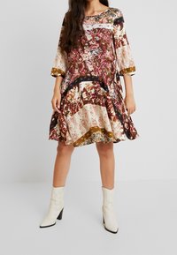 Wielokolorowa sukienka w kwiaty z rękawami 3/4, rozkloszowanym dołem oraz kontrastującymi czarnymi i złotymi akcentami. Noszona z białymi botkami na obcasie.
