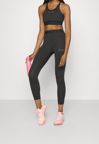 Un reggiseno sportivo nero e leggings a vita alta, entrambi con un logo discreto. Sneakers rosa e una bottiglia d'acqua rosa completano l'outfit.