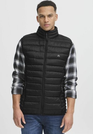Chaleco acolchado negro con cuello alto, cierre de cremallera y diseño de acolchado vertical. Lleva puesto sobre una camisa de cuadros gris, con mangas largas y puños remangados.