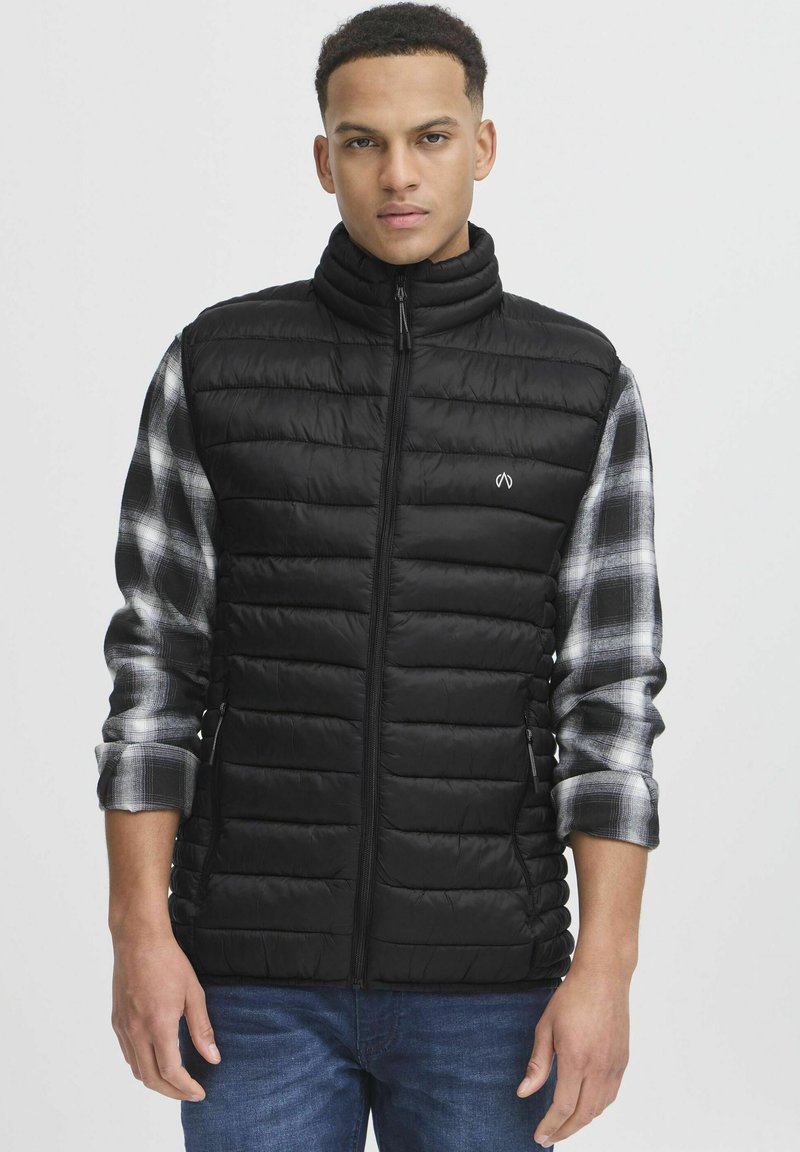 Gilet imbottito nero con colletto alto, chiusura zip e design a costine verticali. Indossato sopra una camicia grigia a quadri, con maniche lunghe e polsini risvoltati.