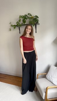 Rød, en-skulder, rynket topp kombinert med løse, svarte bukser. Tregulv, lyse vegger, potteplante på hylle i bakgrunnen, og en nøytral lenestol.