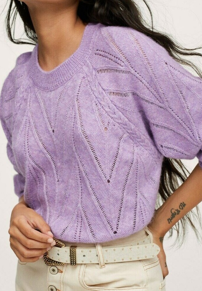 Femme portant un pull en tricot violet clair avec un motif chevron, un pantalon beige et une ceinture beige cloutée, avec un tatouage visible au poignet.