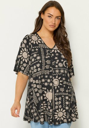 Kvinde med langt hår iført sort tunika med beige sol-, måne-, stjerne- og blomsterprint, kombineret med lyseblå jeans.