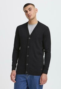 Schwarzer cardigan aus weichem Strickmaterial mit V-Ausschnitt, sechs Knöpfen auf der Vorderseite und gerippten Bündchen. Über einem grauen Hemd getragen, kombiniert mit blauen Jeans.