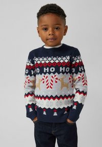 Młody chłopiec ubrany w granatowy sweter świąteczny z napisem "HO HO HO", wzorami cukierków, reniferów i płatków śniegu, stojący z rękami w kieszeniach.