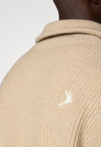 Maglione lavorato a maglia beige con un design a coste testurizzato. Presenta un logo di un uccello ricamato in bianco sulla spalla sinistra. Dettaglio del colletto piegato.