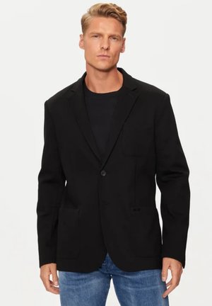 Blazer nero con chiusura a un bottone, due tasche frontali e un tessuto texturizzato. Indossato sopra una camicia nera e jeans blu.