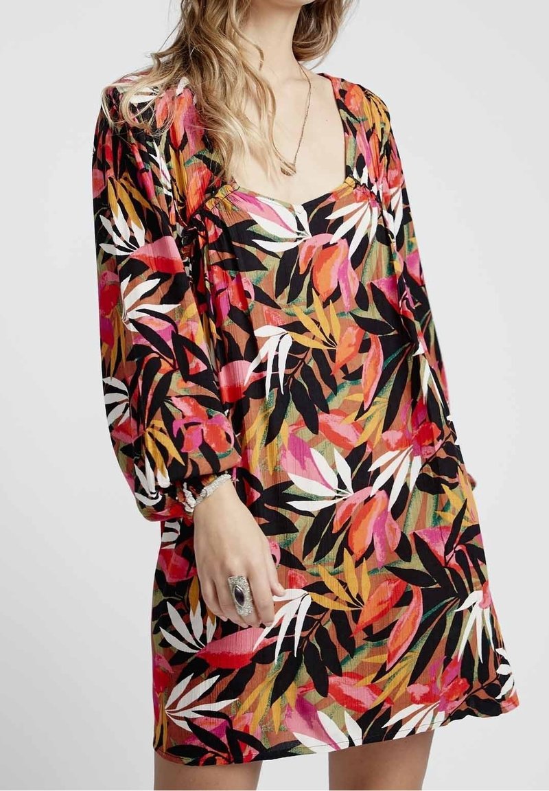 Femme portant une robe à manches longues avec un motif coloré de feuilles tropicales dans des nuances de rose, orange, noir, blanc et vert.