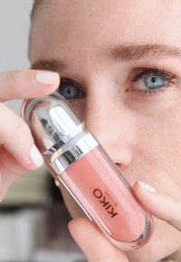 Bouteille en verre opaque avec un capuchon argenté, contenant un liquide de teinte pêche. Nom de la marque "KIKO" imprimé sur le côté. Forme lisse et arrondie.