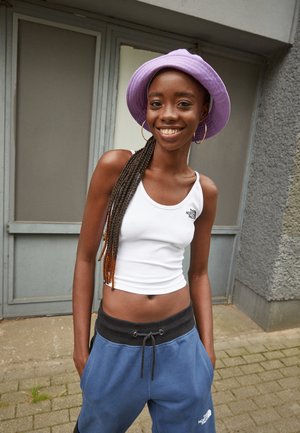 Smilende kvinde med lange fletninger bærer en lilla bucket hat, hvid cropped tanktop, blå sweatpants og står udendørs på belagt grund.