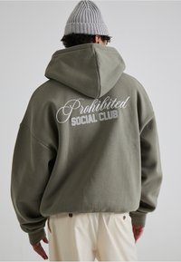 Sweat à capuche oversize vert olive avec un grand texte brodé blanc "Prohibited Social Club" au dos, muni d'une capuche avec cordon de serrage et de poignets côtelés.