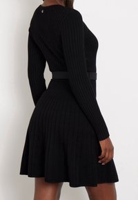 Robe en maille côtelée noire avec manches longues, corsage ajusté, jupe évasée, et une ceinture noire à la taille ; dotée d'un col rond.