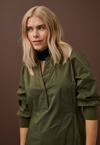 Plavuša u bluzama maslinasto zelene boje s naboranim rukavima gleda u stranu na smeđoj pozadini.