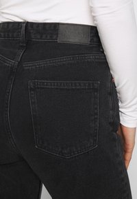 Vista trasera de una persona que lleva puestas jeans de mezclilla negras con una etiqueta de la marca Monki y una camisa blanca de manga larga.