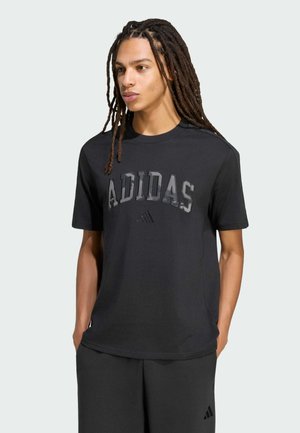 Giovane con lunghi dreadlocks che indossa una t-shirt nera Adidas e pantaloncini neri, in piedi con le mani in tasca, che guarda di lato.